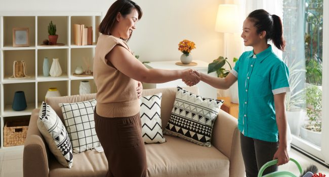 Satisfied Asian woman shaking hands with cleaning service worker to thank her for her work while they standing in living room
